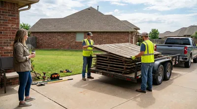 Construction workers in high-visibility vests loading weathered wooden fence panels onto a utility trailer in a residential driveway, with brick homes and green lawns visible in the background.