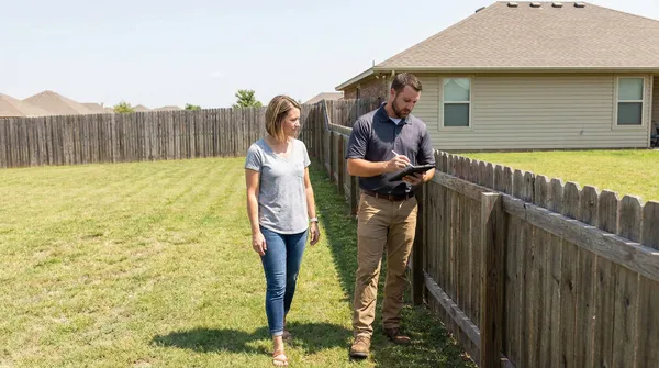 Professional fence contractor inspecting and measuring an existing wooden fence in a residential backyard while discussing options with a homeowner on a sunny day.