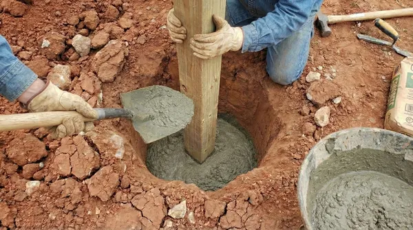 Fence post installation in red clay soil showing multiple concrete-filled post holes, with work gloves gripping a wooden post being set into position.